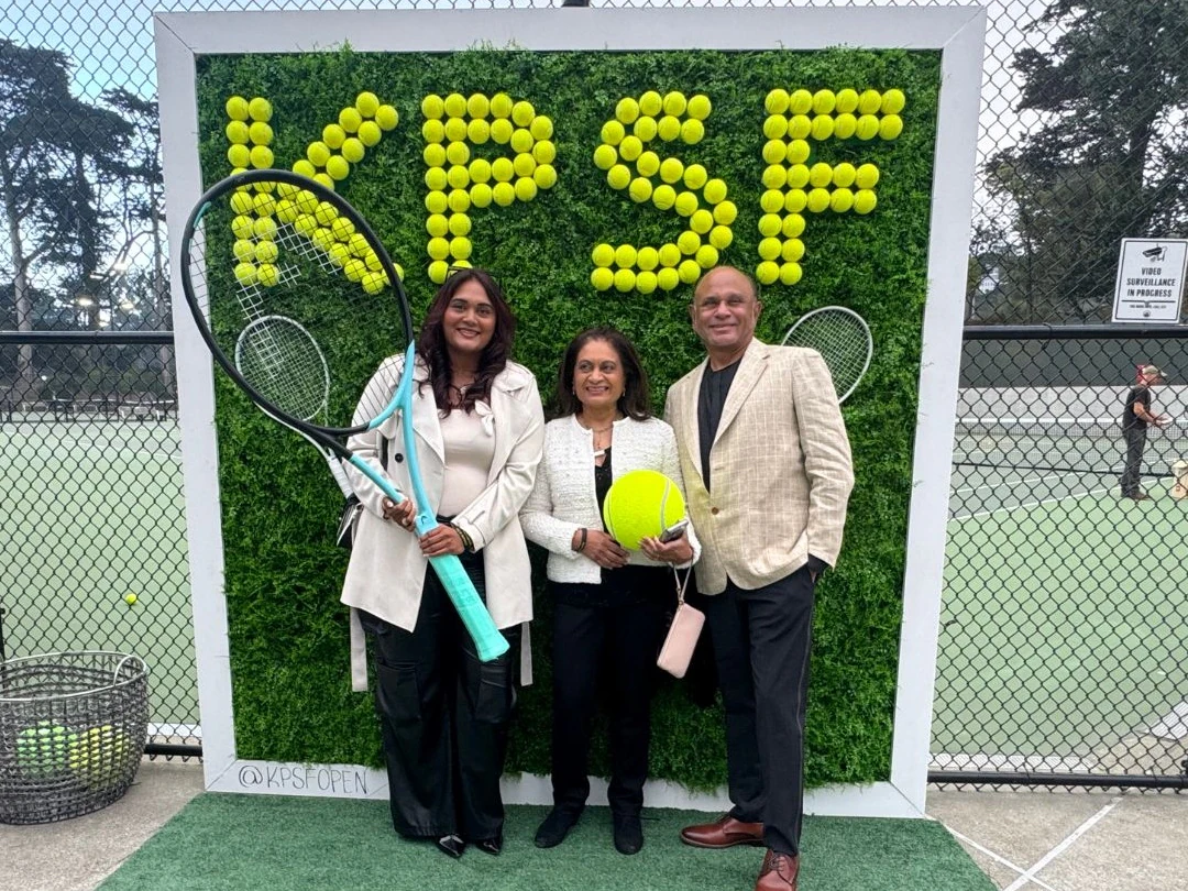 Guests photographed at a branded greenery wall spelling KPSF in tennis balls, beside the courts at a tennis open