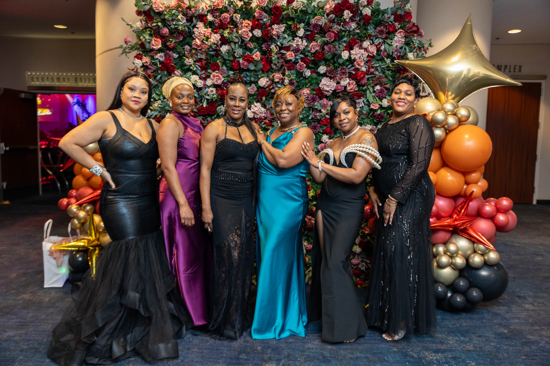 Guests in evening gowns photographed together at a jewel-toned flower wall in a Queens banquet hall