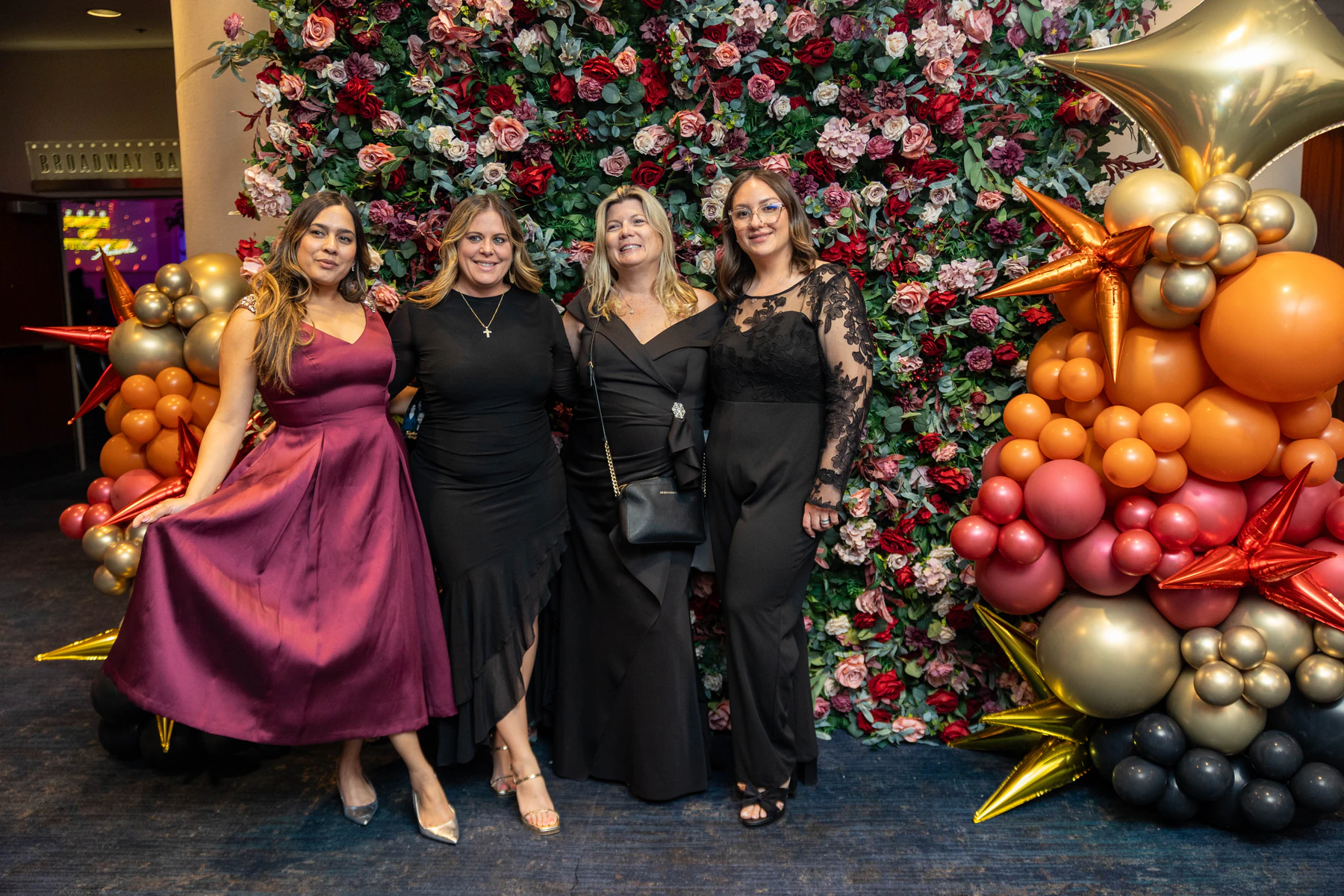 Four guests in evening dress photographed in front of a jewel-toned flower wall and balloon installation at a New York gala