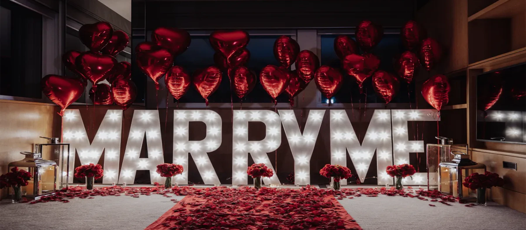 Marriage proposal setup in a New York apartment with illuminated MARRY ME letters, red heart balloons and rose petals
