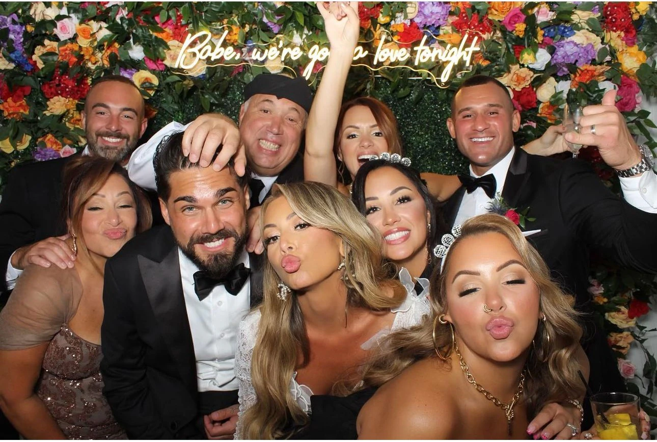 Bride, groom and their wedding party photographed together in front of a bright floral wall with a custom neon sign at a New York wedding