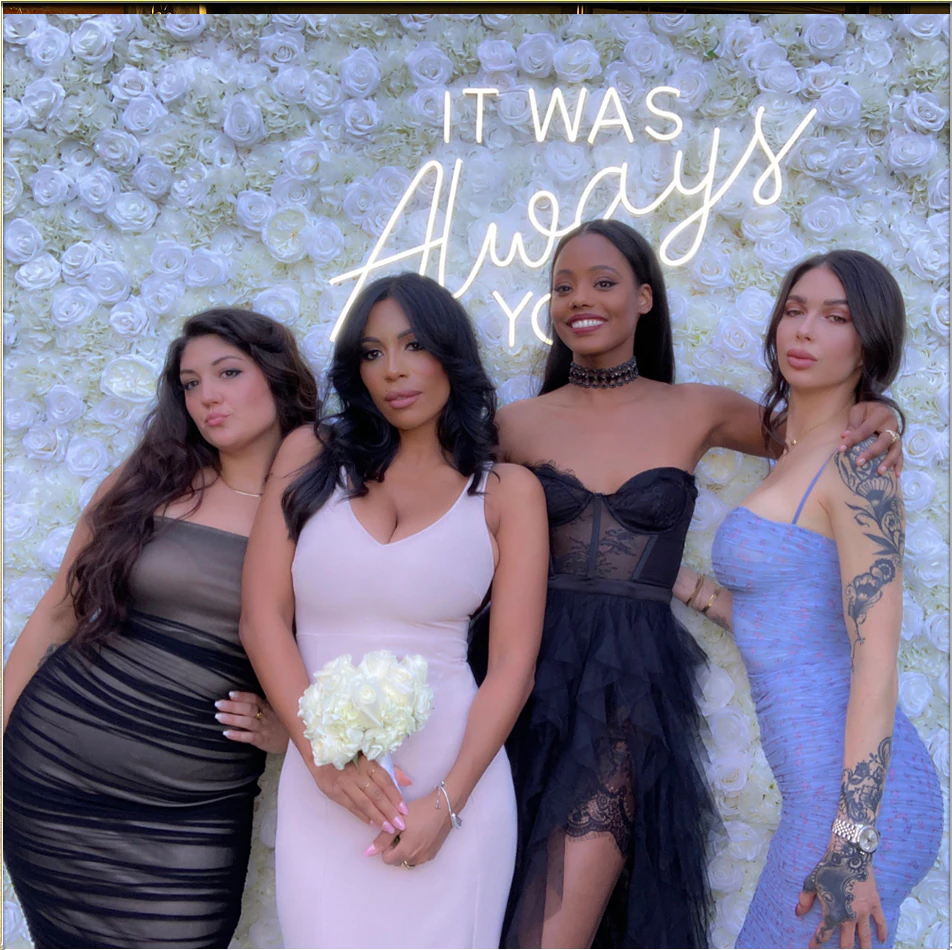 Guests photographed in front of a white rose flower wall with a custom neon sign at a wedding